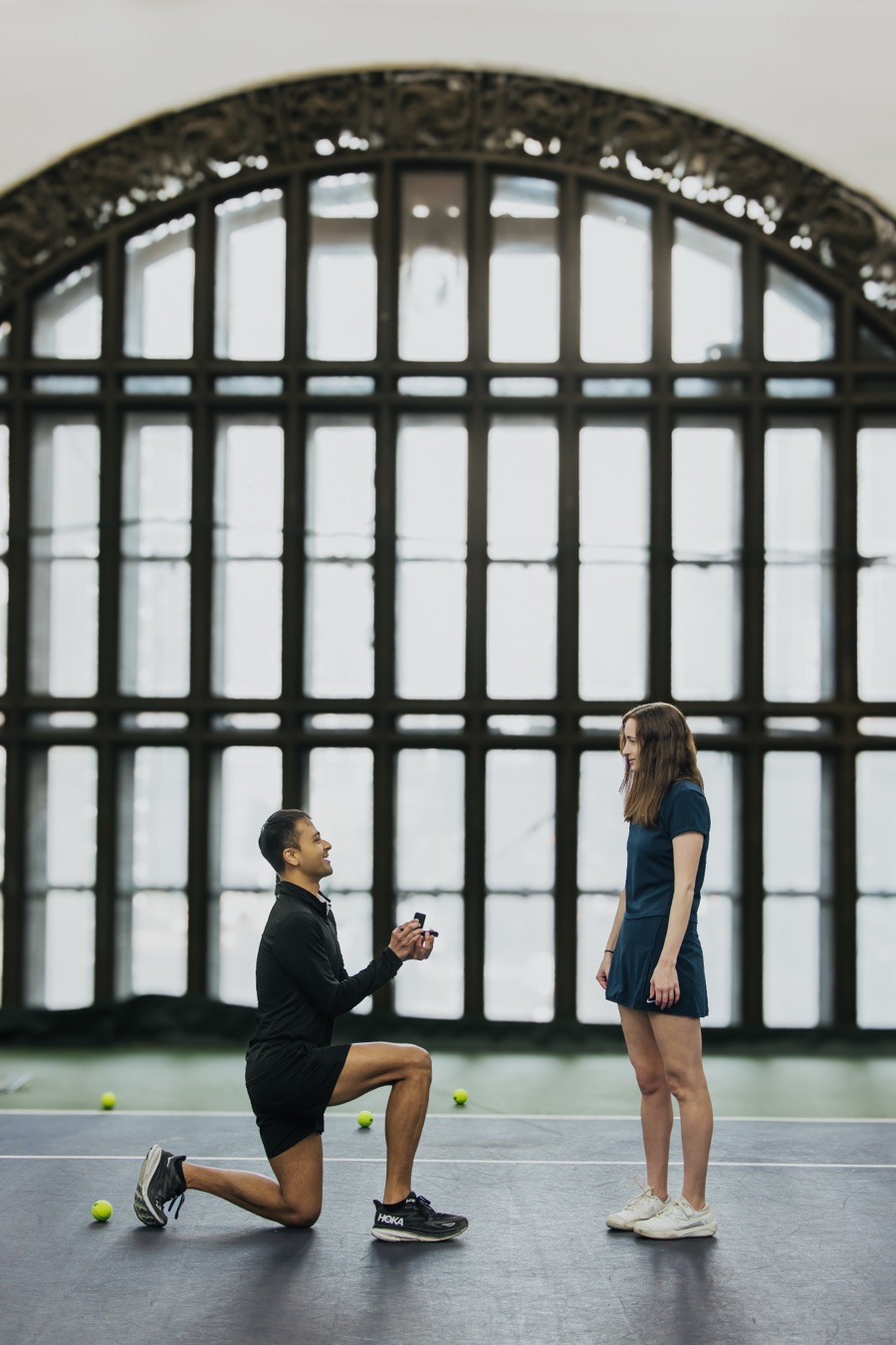 Shayon on one knee proposing to Amanda on a tennis court beneath a tall arched window